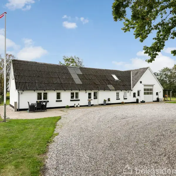 Lang, hvid bygning med sort tag står på grusplads omgivet af grønne træer og græsplæne. En flagstang med dansk flag ses ved siden af bygningen.