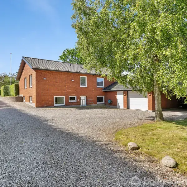 Et rødt murstenshus med hældningstag står stille ved en grusvej. Omkranset af grønne hække og træer, med en garage til højre, skaber det en idyllisk landejendommens atmosfære.