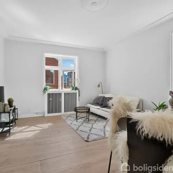 Stuen har en sofa mod en hvid væg, ved siden af et lille bord og lampe. På gulvet ligger et tæppe. Der er store vinduer, som giver rummet lys. Billedet indeholder teksten "boligsiden".