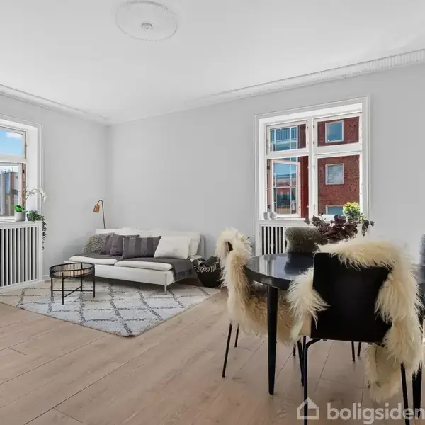 En lys stue med en sofa og et sofabord på et tæppe. En spiseplads med fire stole dækket af skind ved et rundt bord. Store vinduer slipper sollys ind.