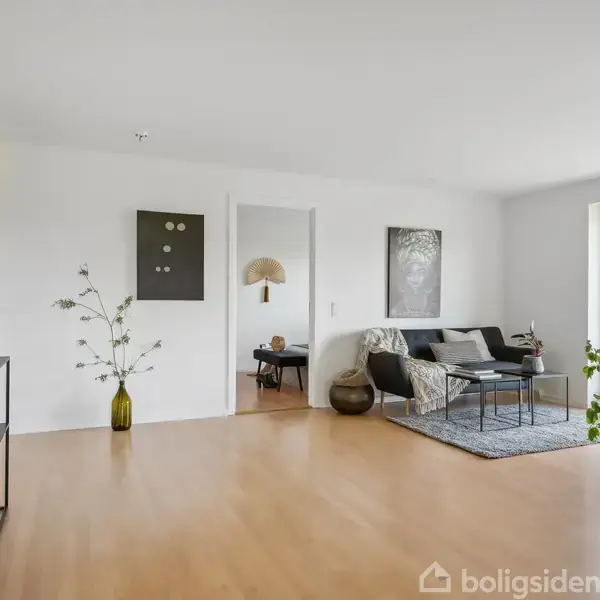 Stue med minimalistisk indretning; en sofa og bord står på et tæppe ved vinduet. En sort reol med dekorationer står til venstre, og planter skaber et grønt miljø.