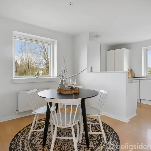 Et rundt spisebord med tre stole står på et mønstret tæppe i et lyst køkkenalrum. To store vinduer giver udsigt til et grønt landskab udenfor.