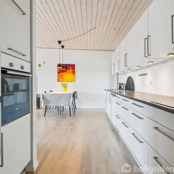 Moderne køkken med hvide skabe og sort bordplade. Køkkenet fører til en spisestue med et bord og stole. Træloft og en farverig maleri på væggen.