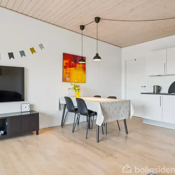 Fladskærms-tv står stationært på sort møbel. Til højre er et spisebord med fire stole, belyst af hængelampe. Køkkenet har hvide skabe. Loftet er af lyst træ.