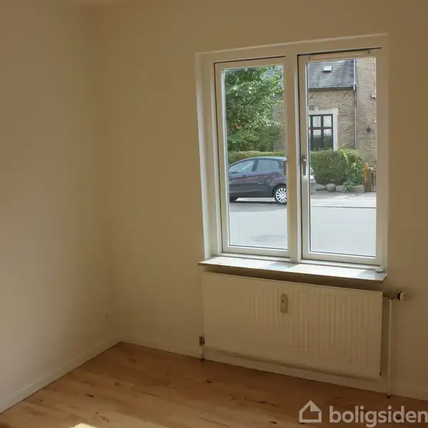 Et lyst, tomt værelse med trægulv indeholder et vindue med udsigt til en gade, hvor en bil er parkeret. Vindueskarmen har en radiator nedenunder.