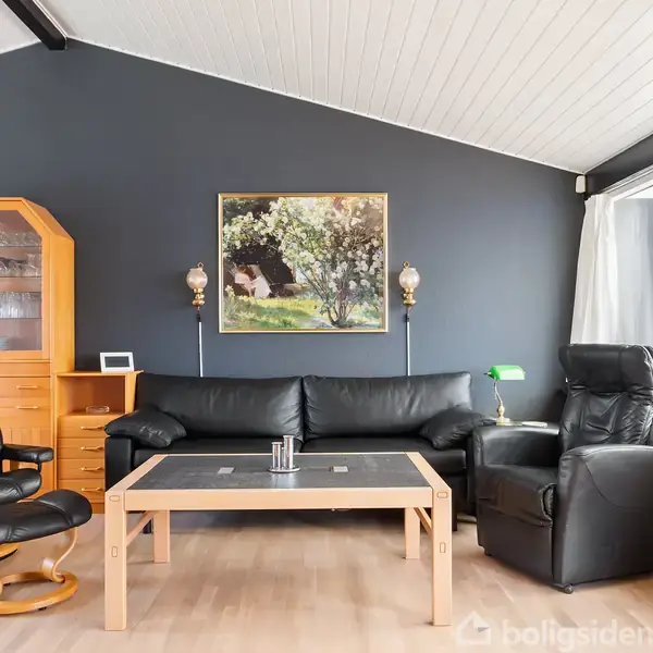 Sort lædersofa og lænestole står omkring et trækaffebord. I baggrunden er en mørk væg med et maleri og to væglamper. Ved siden står et skab og store vinduer.
