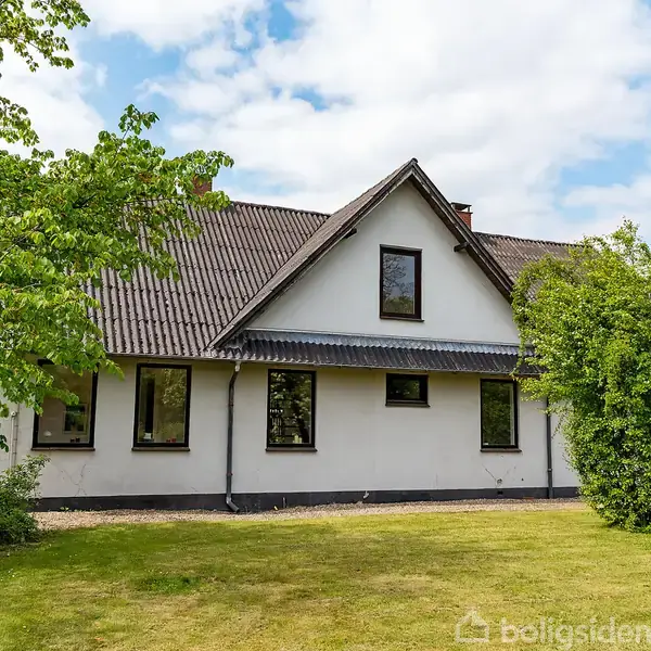 Et hvidt hus med gråt tag står stille i en grøn have, omgivet af træer og græsplæne under en blå himmel med skyer.