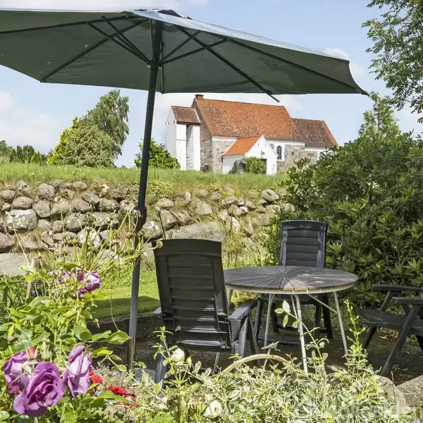 Et bord med fire stole står under en stor parasol. Blomster omgiver møblerne, og i baggrunden ses en bygning med rødt tag bag en stengærde.