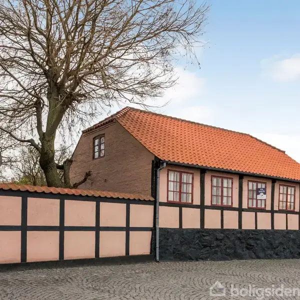 Bindingsværkshus står stille, omgivet af brostensbelagt gårdsplads. Huset har rødt tegltag, malede paneler og et træ, der rækker op imod himlen i baggrunden.