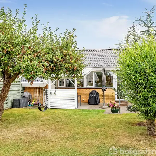 Huset med gråt tag står i baghaven omgivet af grønne træer og en velplejet græsplæne. En gynge hænger fra et træ, og der er en grill foran huset.