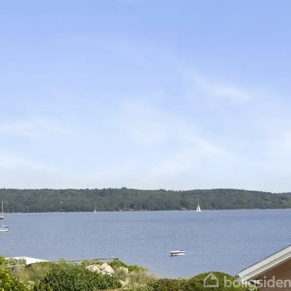 Hav udsigt med sejlbåde på vandet, omgivet af skov på den fjerne bred. Huse med røde tage og grønne buske i forgrunden.