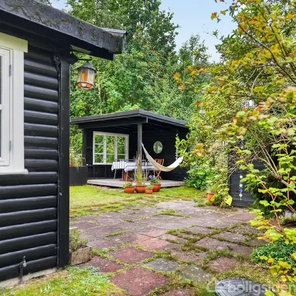 Sortmalet havehus med terrasse og hængekøje. Terrassen har et bord med dug og stole. Omgivet af frodig have med buske og træer. Ingen tekst synlig.
