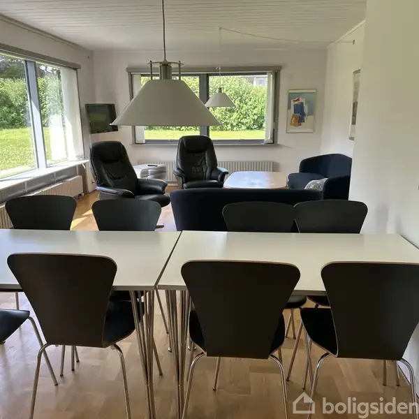 Et spisebord omgivet af sorte stole står i en stue med store vinduer mod en grøn have. I baggrunden ses lænestole og en sofa under en moderne loftslampe.