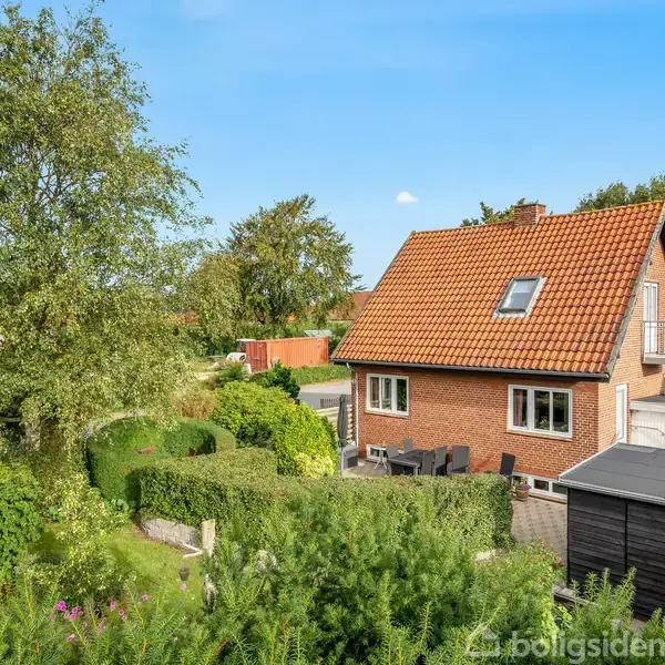 Rødt murstenshus med skråt tag ligger stille i en velplejet have med buske og træer. Solrig terrasse ved siden af garagen. Blå himmel i baggrunden.