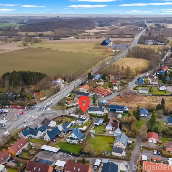 Rødt lokationsikon markerer en ejendom blandt rækkehuse og grønne områder; veje og marker strækker sig i baggrunden.