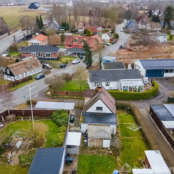 Luftfoto af et boligområde viser huse med haver og garager. En snoet vej skærer gennem området, omgivet af træer og marker i baggrunden.