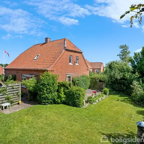 Rødt murstenshus med rødt tegltag står i en velplejet have. Ingen mennesker er synlige. Området omkring er grønt, med buske og træer, og en blå himmel med skyer ses ovenover.
