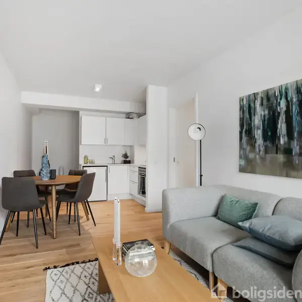 En stue med en grå sofa, dekoreret med puder, står i forgrunden. Et lille sofabord med lys foran, mens et spisebord med stole ses ved det åbne køkken i baggrunden.