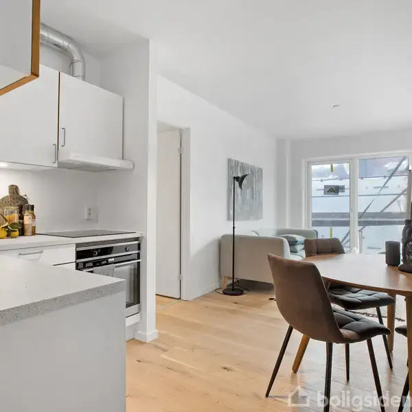 Køkken og stue i ét åbent rum; køkkenet har hvide skabe og moderne apparater. Stuen har træbord med stole samt sofa i lysdagt belyst af stort vindue.