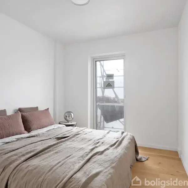 En dobbeltseng med beige sengetøj står roligt i et lyst soveværelse. Ved siden af er et lille rundt natbord med en lampe. Et vindue skaber naturligt lysindfald. Ingen tekst.