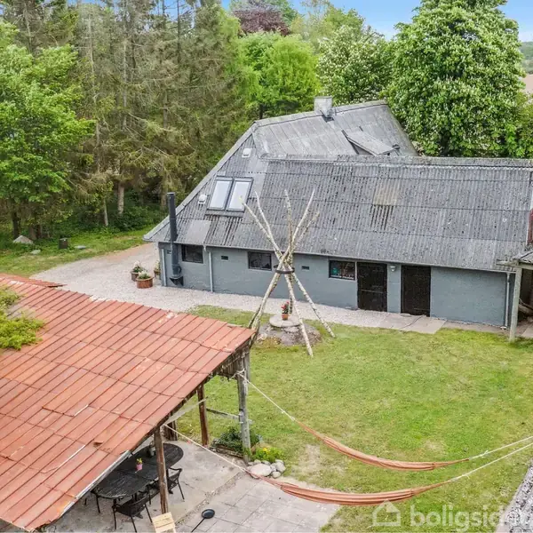En grå, traditionel bygning står i en grøn have med træer i baggrunden. Bygningen har et skråt tag med ovenlysvindue. Foran ses et overdækket område med rødt tag.