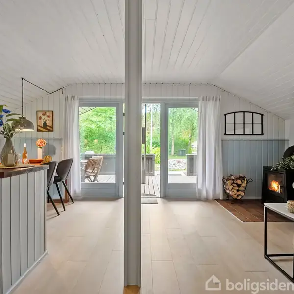Rummet viser en stue med pejs, sofabord og sofa til højre. En spiseplads med stole er forbundet til køkkenet til venstre. Store vinduer åbner mod en terrasse og have.