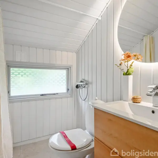Et moderne badeværelse har hvidt træpaneler. En rund oplyst spejl hænger over en vask med træskab. Toilet står ved siden af, ved et matteret vindue. Blomstervase pryder vasken.