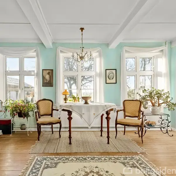 Et lyst værelse med træmøbler og planter, dekoreret med malerier. To lænestole står ved et bord under en lysekrone. Store vinduer giver udsigt til en frodig have.