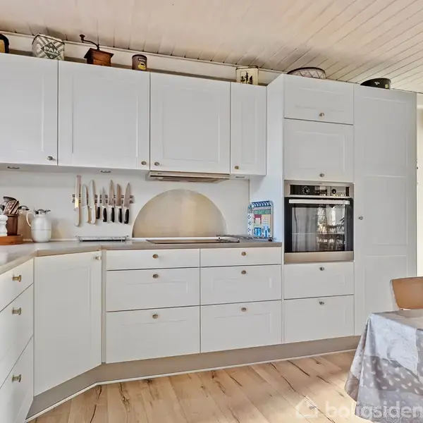 Hvidt køkken med skabsfacader og indbygget ovn. Bordpladerne har køkkenredskaber og knivholder. Et rundt lysindfald er synligt, og der står en dækket stol ved et bord i forgrunden.