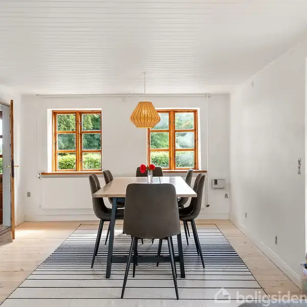 Spisebord med seks stole står på et stribet gulvtæppe. En trælampe hænger over bordet. Rummet har lyse vægge, trægulv og to vinduer med udsigt til grønne omgivelser.