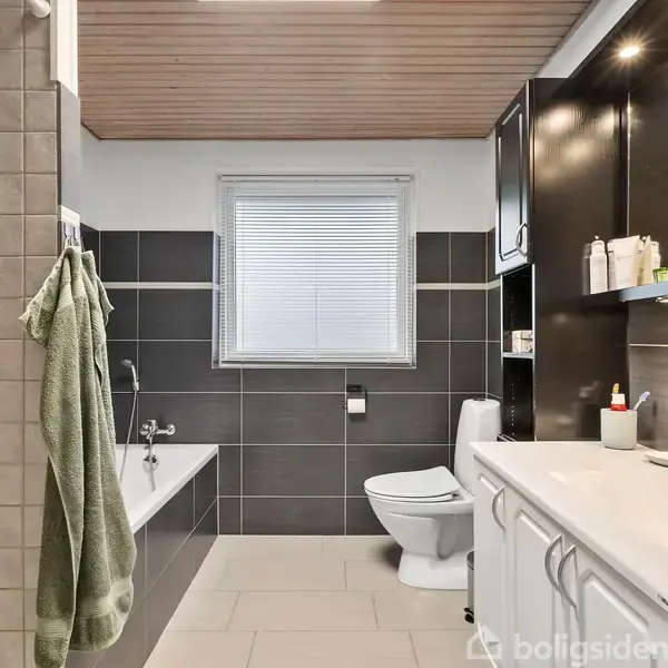 Badeværelse med badekar til venstre, håndklæde hængende, toilet i midten, og vask til højre med spejl. Lyse fliser, mørke skabe, og et vindue med persienner.