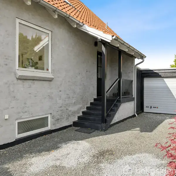Trappe fører op til hus med gråt pudset facade og rødt tegltag. I baggrunden ses en garage med grå port og grusbelagt indkørsel omgivet af buske.