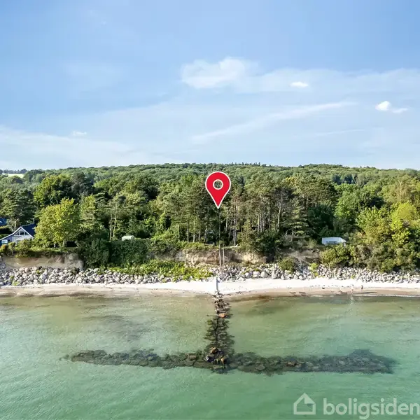 En rød markør viser placeringen i en skov, der grænser op til en strandkant med klart vand og stenbarriere. I baggrunden ses frodig skov og enkelte huse.