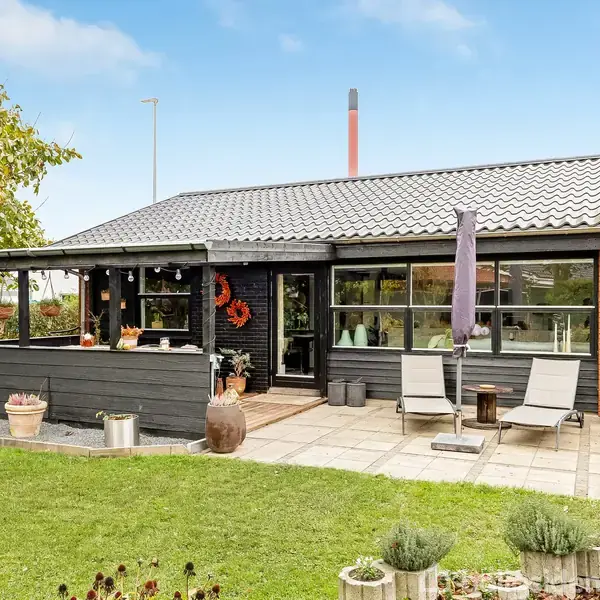 Et hyggeligt hus med en rummelig terrasse er omgivet af en grøn have. To solstole og en parasol er placeret på terrassen, mens planter dekorerer området.