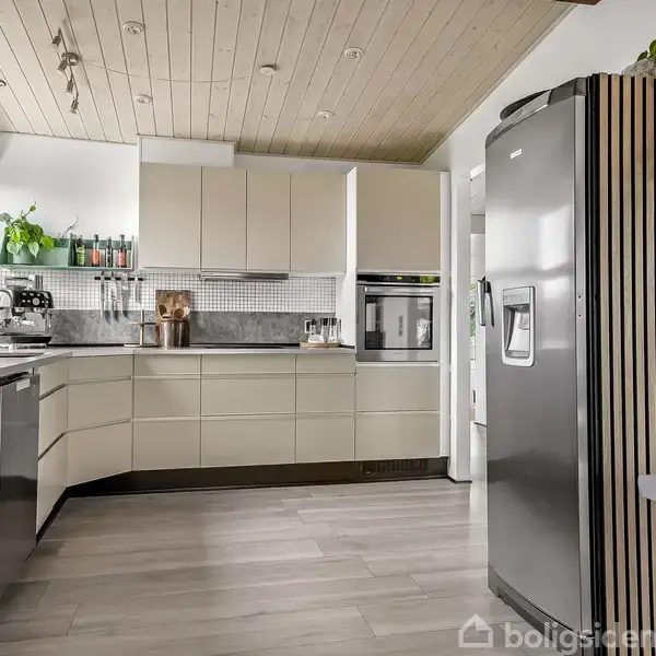 Et moderne køkken med lyse skabe, en integreret ovn, og en stor køleskab i rustfrit stål. Bordpladen er dekoreret med planter. Trægulv og træloft skaber en naturlig atmosfære.