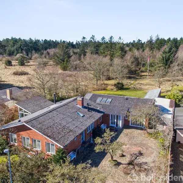 Rødt murstenshus set fra oven omgivet af en stor have med træer. Skovklædt område ses i baggrunden under en klar blå himmel.