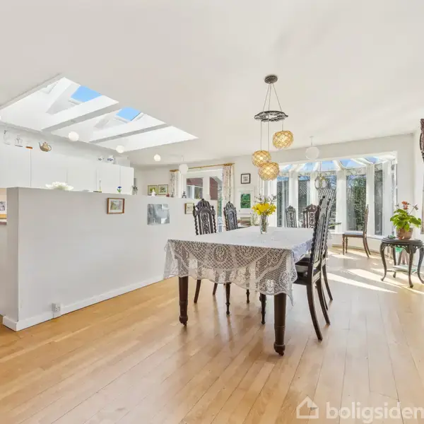 Et lyst køkken-alrum med et spisebord dækket af en dug, omgivet af stole. Køkkenet har hvide skabe, ovenlys og trægulv. Store vinduer slipper masser af naturligt lys ind.