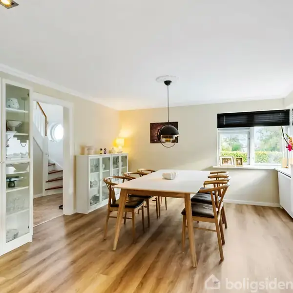 Spisebord med seks stole står i et lyst rum. En pendellampe hænger over bordet. Skabe med glaslåger og vindue med udsigt omgiver området. Ingen tekst findes i billedet.