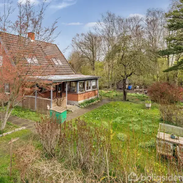 Et rødt murstenshus ligger stille i en frodig have med grønne træer og buske. En terrasse med tag omkranser huset, mens en havebænk står på græsset.