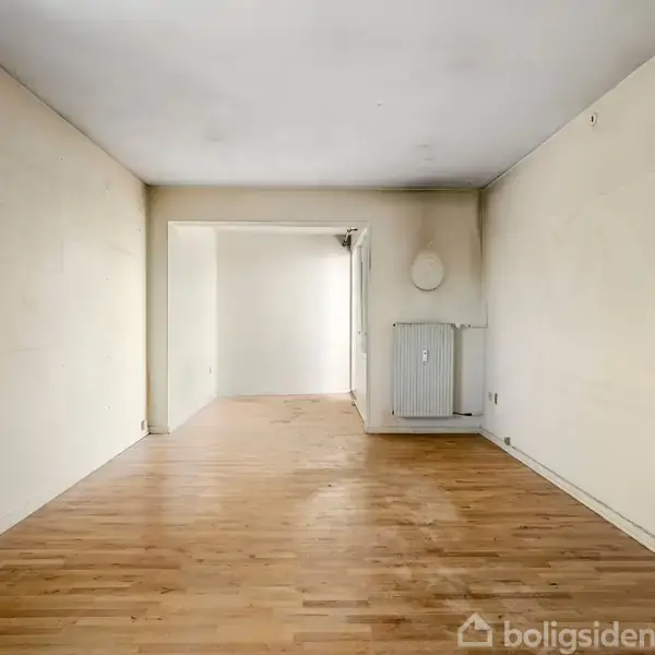 Et tomt rum med trægulv strækker sig ind i en lysere sektion i baggrunden. På væggen er der en radiator, og væggene er lyse.