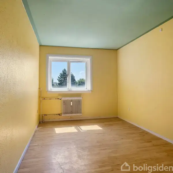 Et tomt rum med lysegule vægge og en lyseblå loft. En radiator står under det store vindue. Dør og træparketgulv er synlige.