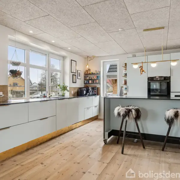 Moderne køkken med lyseblå skabe og sorte bordplader. To barstole med pelssæder står ved en central køkkenø. Store vinduer slipper naturligt lys ind. Plante og dekorationer tilføjer liv.