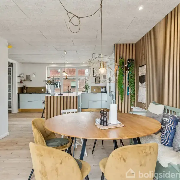 Rundt træbord med gulpolstrede stole står i en moderne spisestue. Køkkenet ses i baggrunden med åbne hylder og planter. Trævægspaneler og lyse gulve skaber en hyggelig atmosfære.