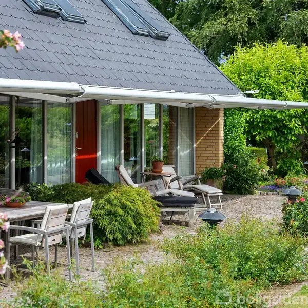 Hus med terrasse og markise, omgivet af grønne buske og blomster. Havemøbler står på flisebelægning. Flere vinduespartier reflekterer lyset. Lyst, frodigt haveområde i baggrunden.