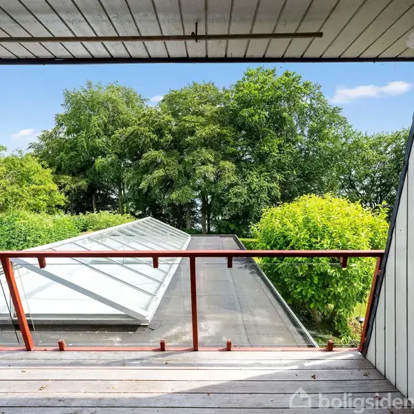 Balkon med trægelænder og udsigt over grøn taghave og frodige træer i baggrunden. Blå himmel kan ses over det grønne landskab.