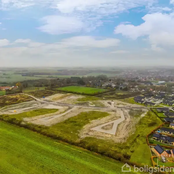 Byggeplads med vejanlæg i et grønt, åbent landskab, under en skyet himmel. I baggrunden ses en by med huse langs vejen.