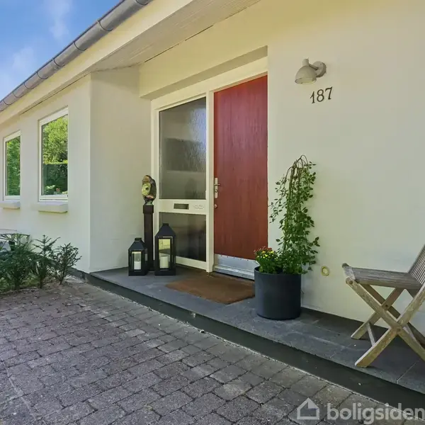 Rød dør pryder husets indgang, flankeret af lanterner og planter. Stol står ved siden af. Husnummer "187" er over døren. Grønt område omgiver huset.