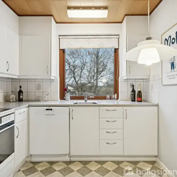 Et hvidt køkken med hvide skabe og apparater. Et vindue med udsigt til træer, en vintagemobil hænger på væggen. Tekst på væggen: "MOR - you will always be with me."