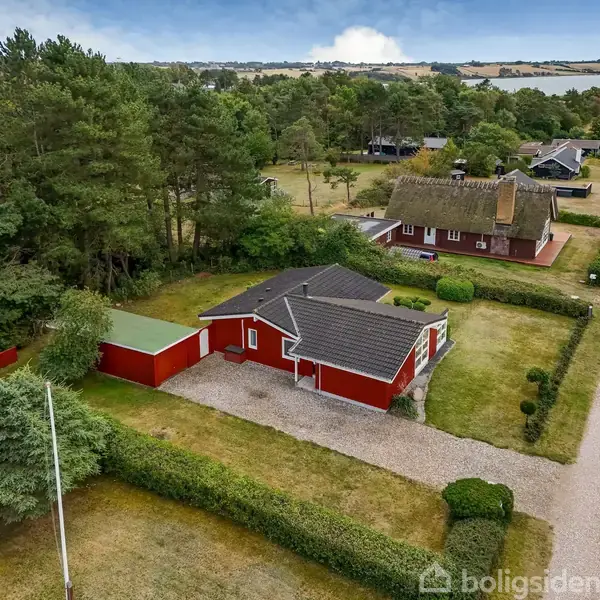 Rødt hus med gråt tag står stille omgivet af træer og en grusvej. Der er en velplejet have og en indkørsel med carport. Udsigt over marker og vand i baggrunden.
