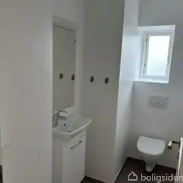 Håndvask under spejl med tre kroge på væggen. Badeværelsesmiljø med vindue til venstre og toilet nedenfor. Enkel og lys indretning.